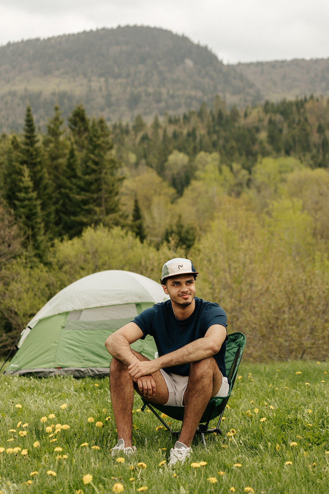 Homme portant une casquette en coton à 5 panneaux de la marque Périple. Modèle colorblock sauge, bleu poudre, marine et crème avec visière brun cacao, assis devant une tente dans un paysage de montagne. Il porte également un t-shirt en laine de mérinos Périple de couleur Marine.