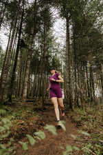 Femme portant un biker short et un t-shirt col V en laine de mérinos pourpre lors d'une course en sentier en forêt.