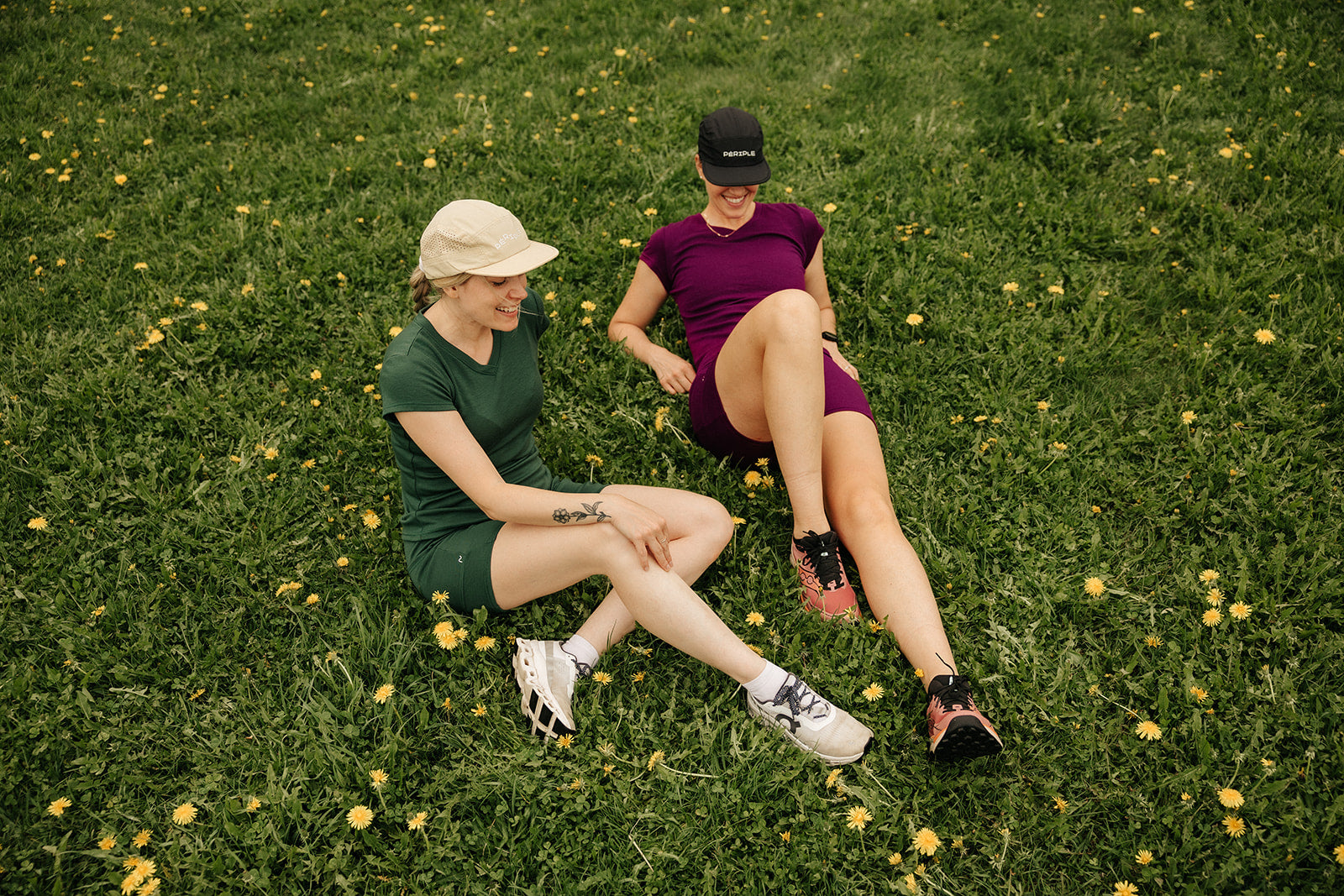Deux femmes portant les casquettes de course Périple beige et noire avec ensembles en laine de mérinos Émeraude et Pourpre.