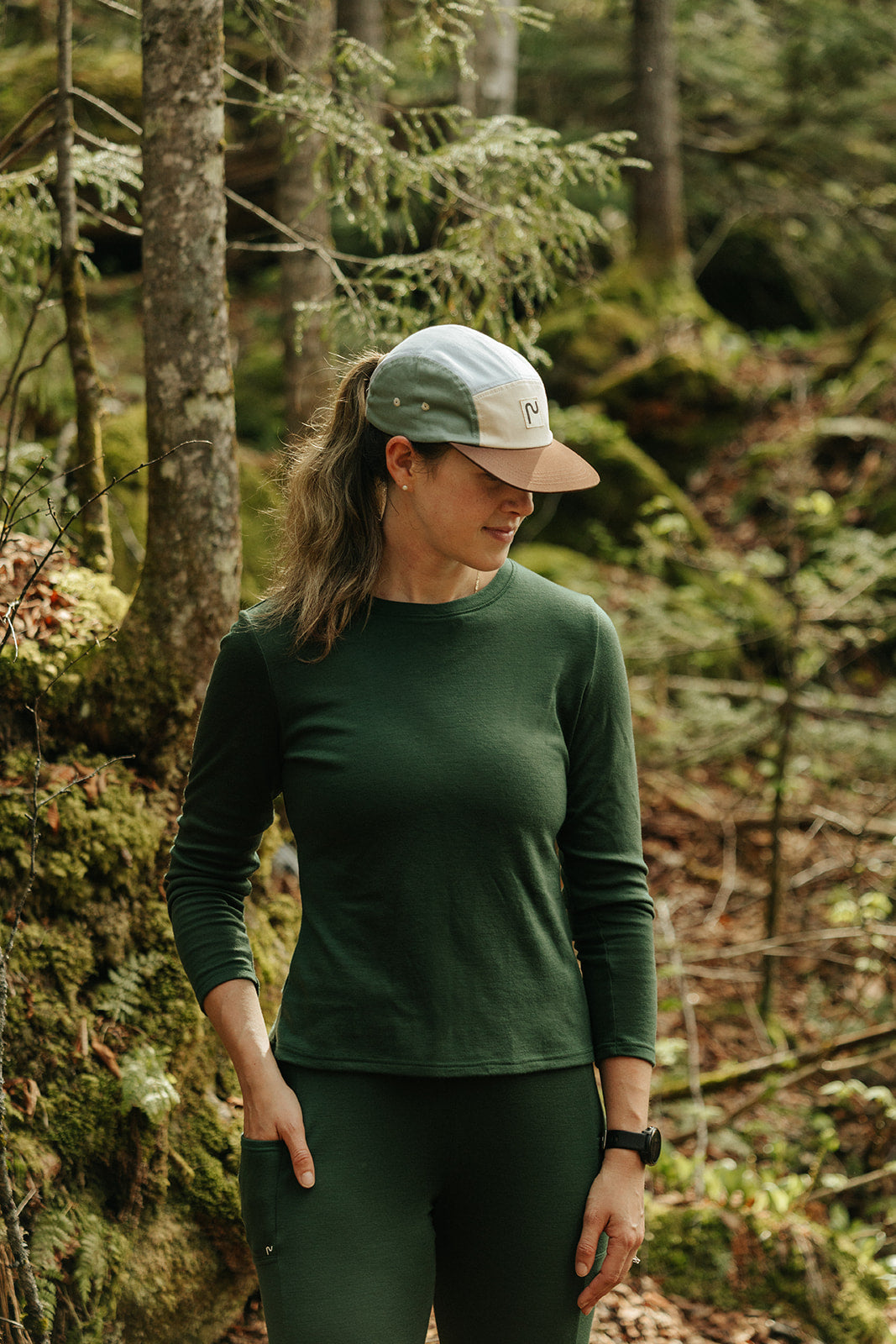 Femme portant un chandail à manches longues et un legging en laine de mérinos Émeraude de Périple lors d'une randonnée en forêt, avec une casquette colorblock lila.