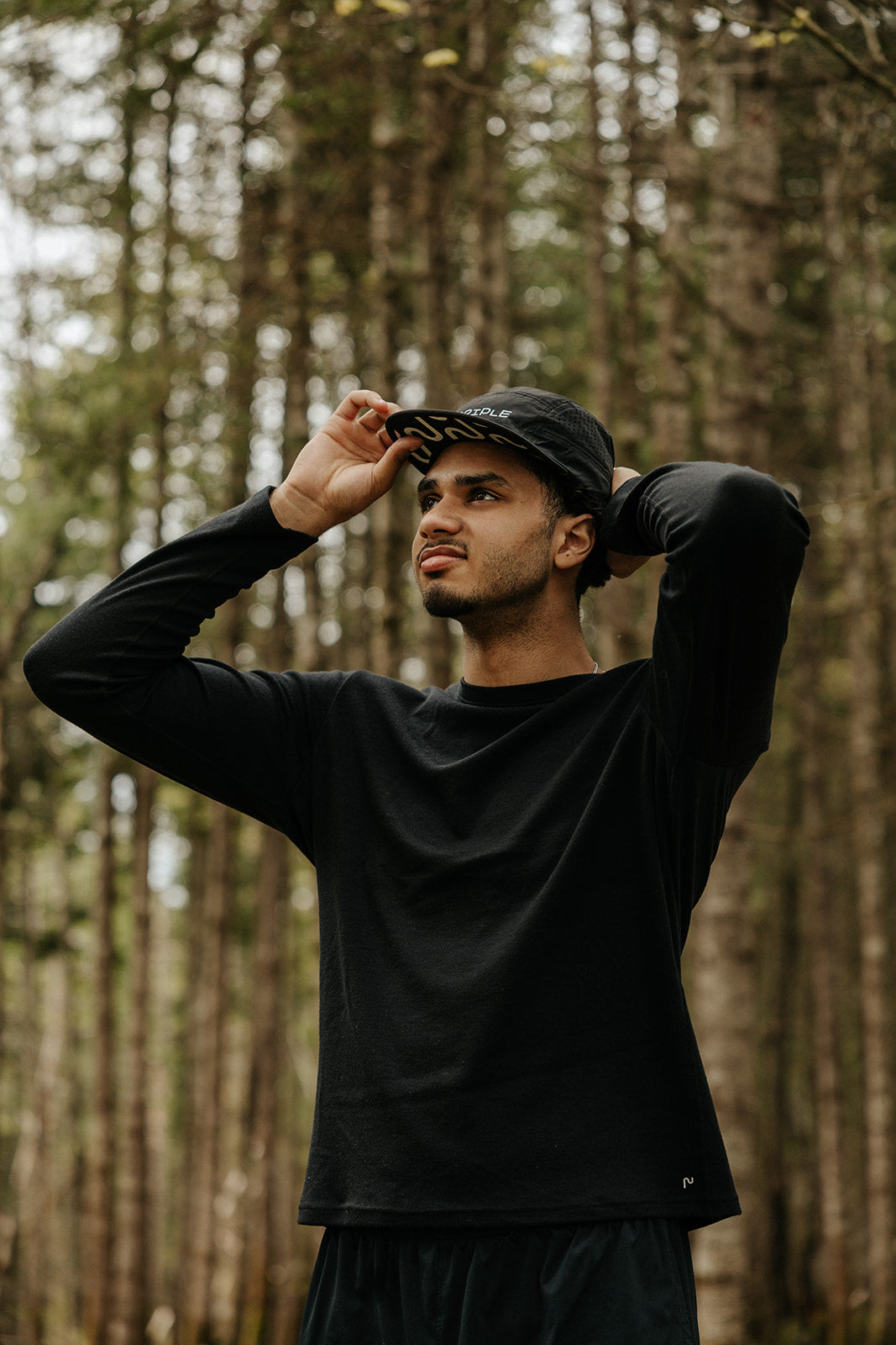 Patrick Déniger portant une casquette de course en nylon noire de la marque Périple et un chandail à manches longues noir en laine de mérinos lors d'une randonnée en forêt.