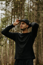 Patrick Déniger portant une casquette de course en nylon noire de la marque Périple et un chandail à manches longues noir en laine de mérinos lors d'une randonnée en forêt.